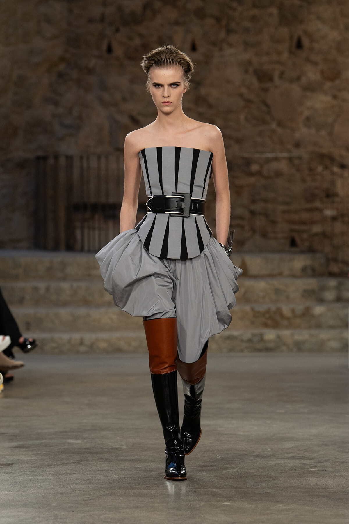 Model on a runway wearing a strapless gray and black striped corset with a voluminous light gray peplum and tall two-tone boots with brown and black panels.