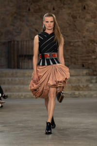 Model walks a fashion runway in a black sleeveless top with white diagonal stripes, a wide red belt, and a voluminous peach draped skirt, carrying a small handbag.