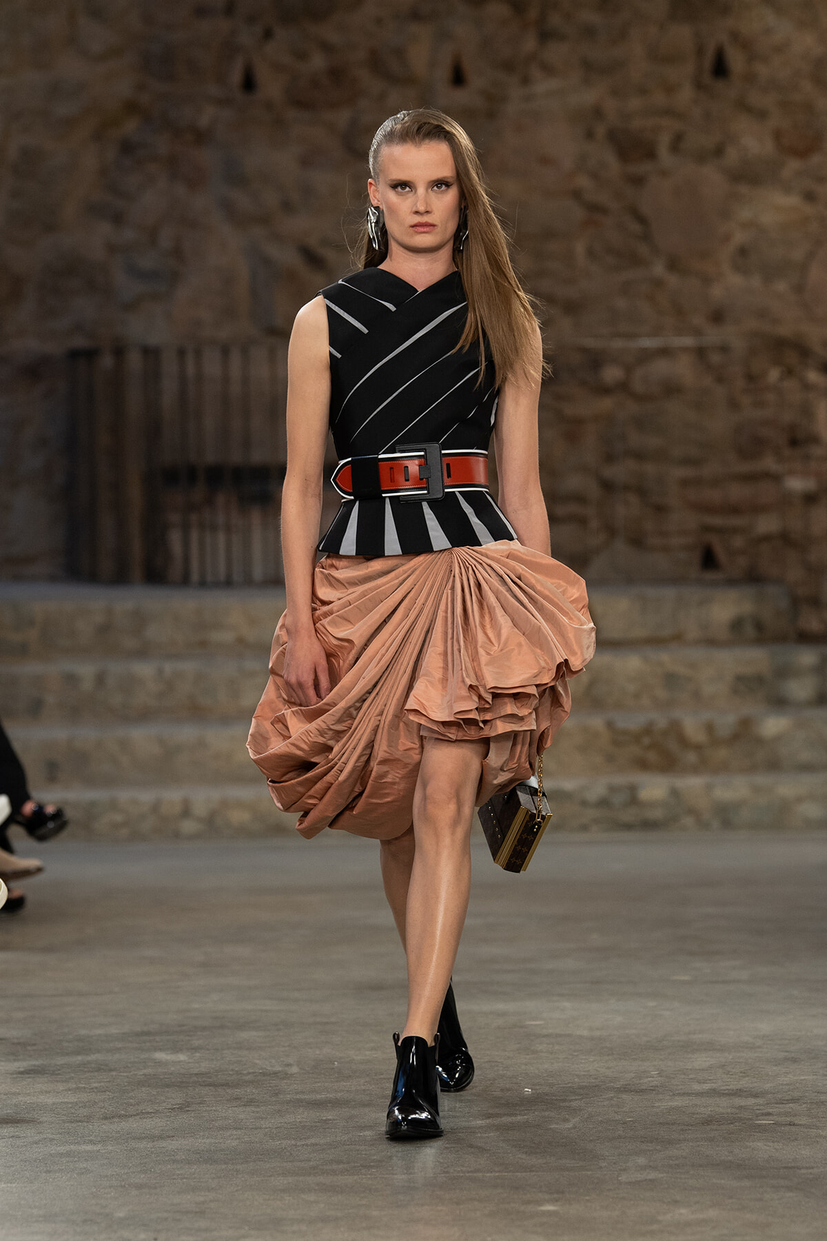 Model walks a fashion runway in a black sleeveless top with white diagonal stripes, a wide red belt, and a voluminous peach draped skirt, carrying a small handbag.