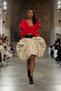 Runway model wearing a bold red sculpted top with a large beige bubble skirt and a wide black belt, walking the catwalk in black ankle boots.