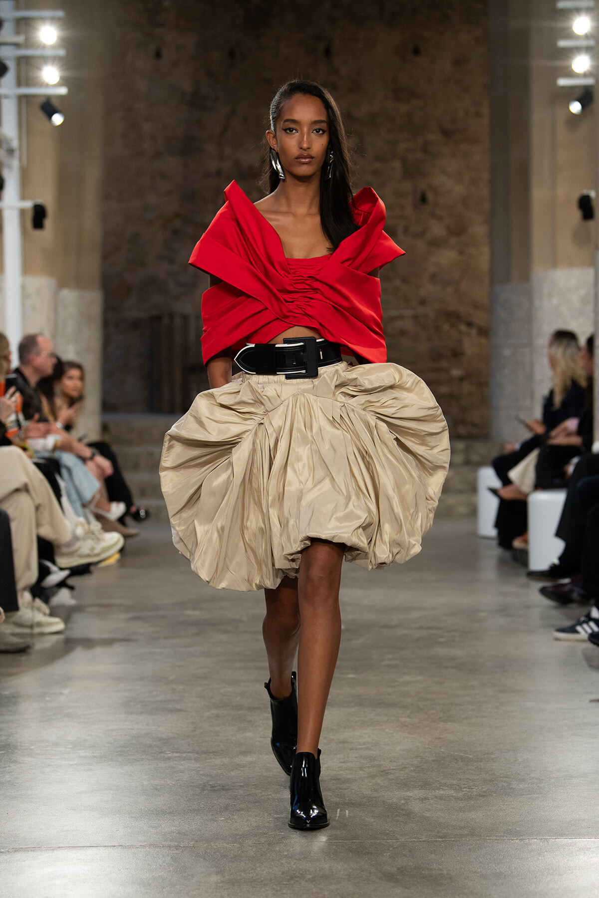 Runway model wearing a bold red sculpted top with a large beige bubble skirt and a wide black belt, walking the catwalk in black ankle boots.