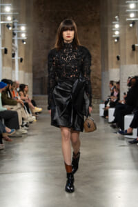 Model walking a fashion runway in a black lace high-neck top and structured leather skirt, carrying a small brown handbag.
