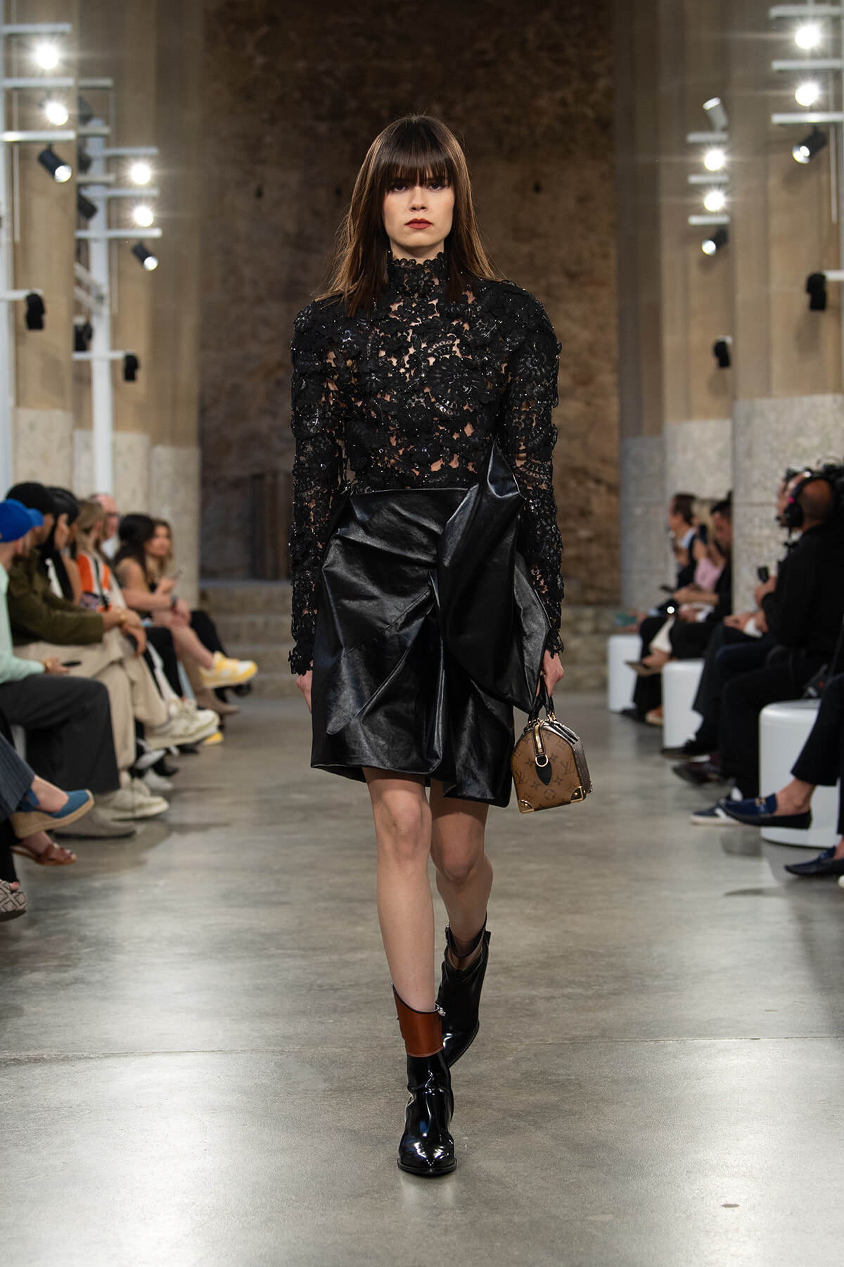 Model walking a fashion runway in a black lace high-neck top and structured leather skirt, carrying a small brown handbag.