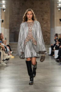 Model walks a fashion show in a silver sequined cape dress with brown bead detailing, paired with black knee-high boots and a small brown handbag.