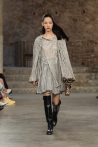 Model walking a runway in a metallic silver patterned dress with a cape, belt, and knee-high black boots.