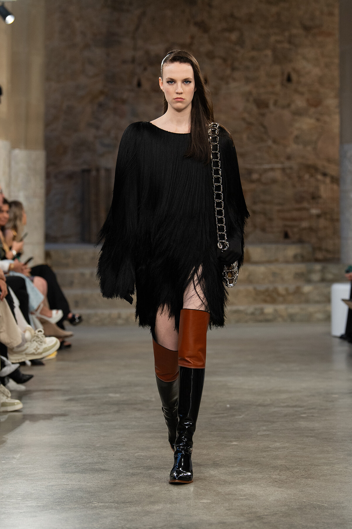 Model walks a runway in a black fringed dress, tall brown-and-black thigh-high boots, and a chunky chain strap bag in an industrial setting.