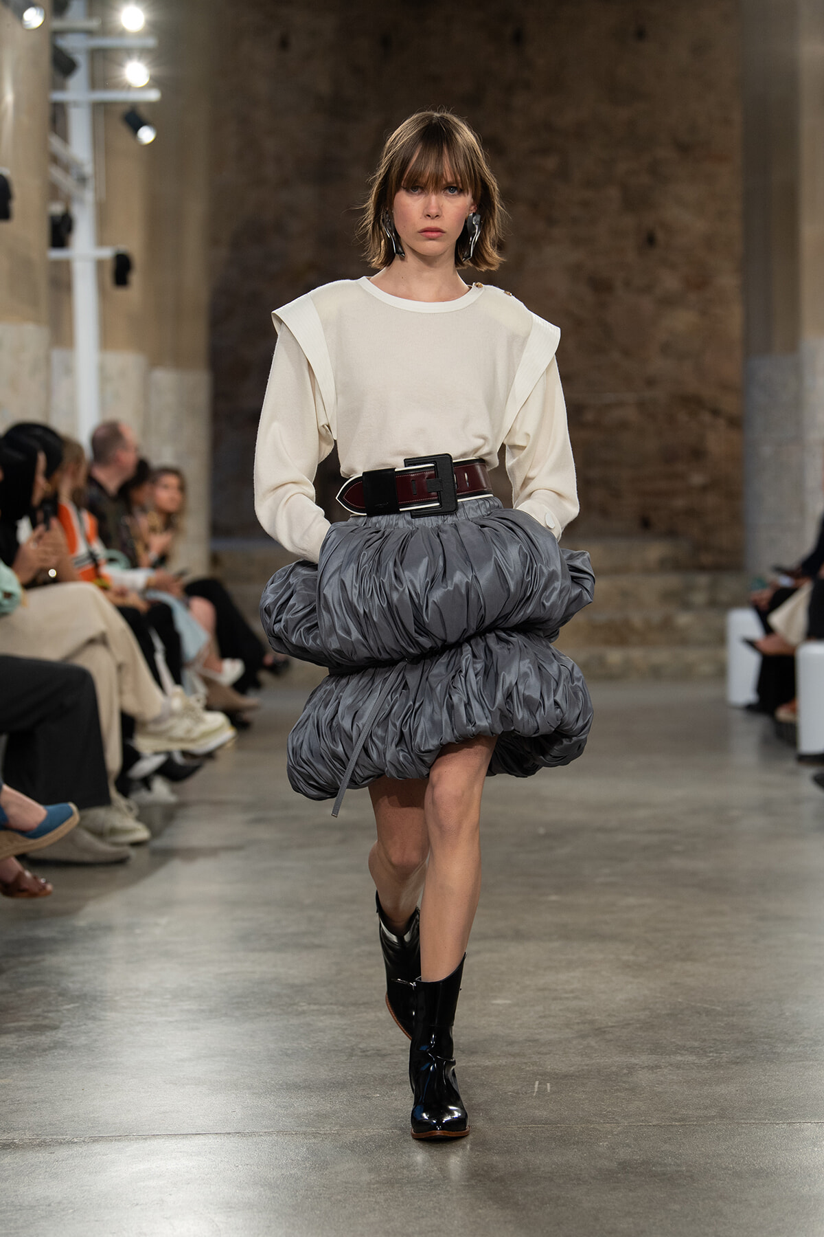 Model on a runway wearing a cream sweater and a voluminous gray bubble skirt with a wide belt and black ankle boots.