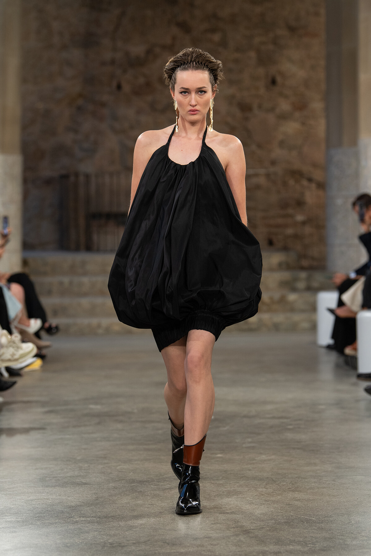 Model walking a runway in a loose black halter bubble dress with gold hoop earrings and glossy black ankle boots, set in an industrial-style venue.