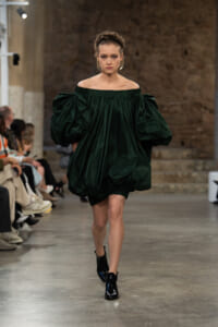 Model walks the runway in an off-the-shoulder emerald green, voluminous dress with oversized puff sleeves.
