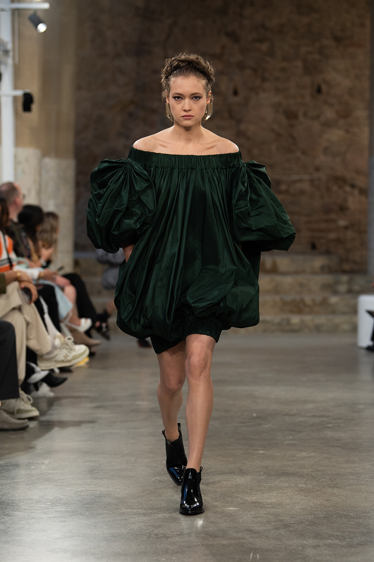 Model walks the runway in an off-the-shoulder emerald green, voluminous dress with oversized puff sleeves.