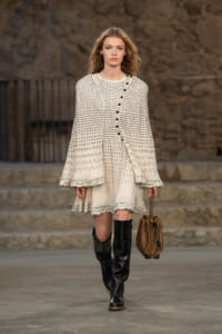 Model walking a runway in a cream dotted dress with a matching cape, black knee-high boots, and a brown quilted handbag with chain strap.