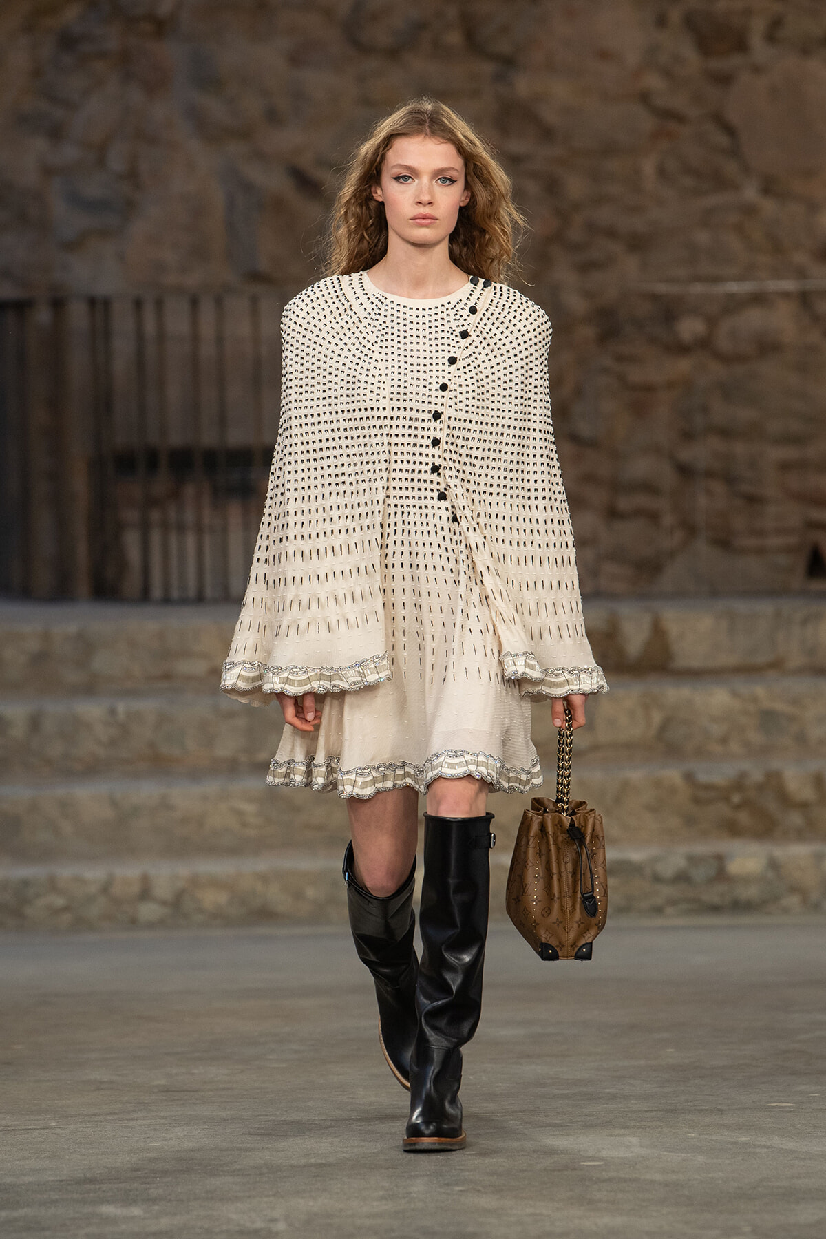 Model walking a runway in a cream dotted dress with a matching cape, black knee-high boots, and a brown quilted handbag with chain strap.