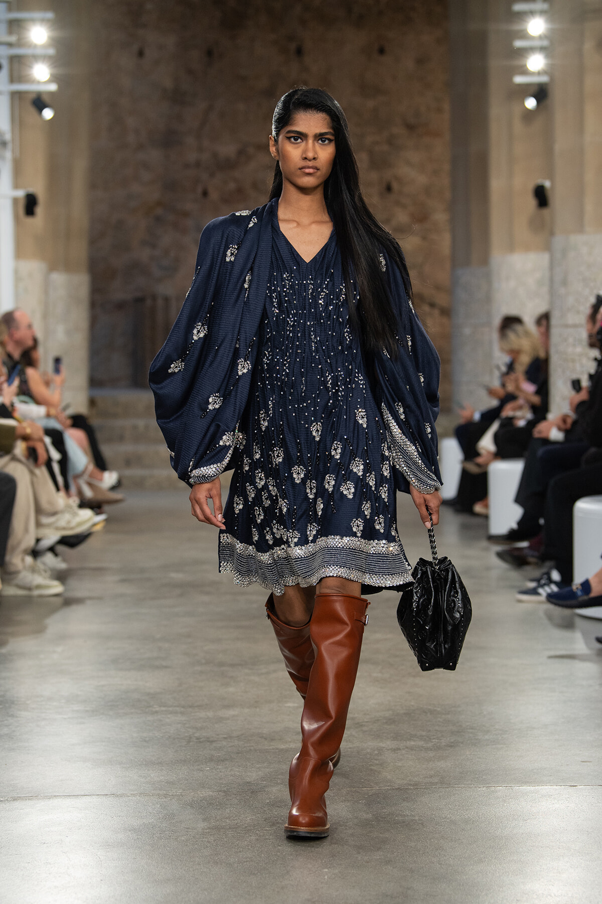 Model walks a fashion runway in a navy embroidered dress with voluminous sleeves and silver embellishments, paired with brown knee-high boots and a black handbag.