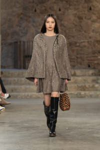 Model walks runway in a sparkling gray cape over a matching dress, paired with black knee-high boots and a checkered brown bag.