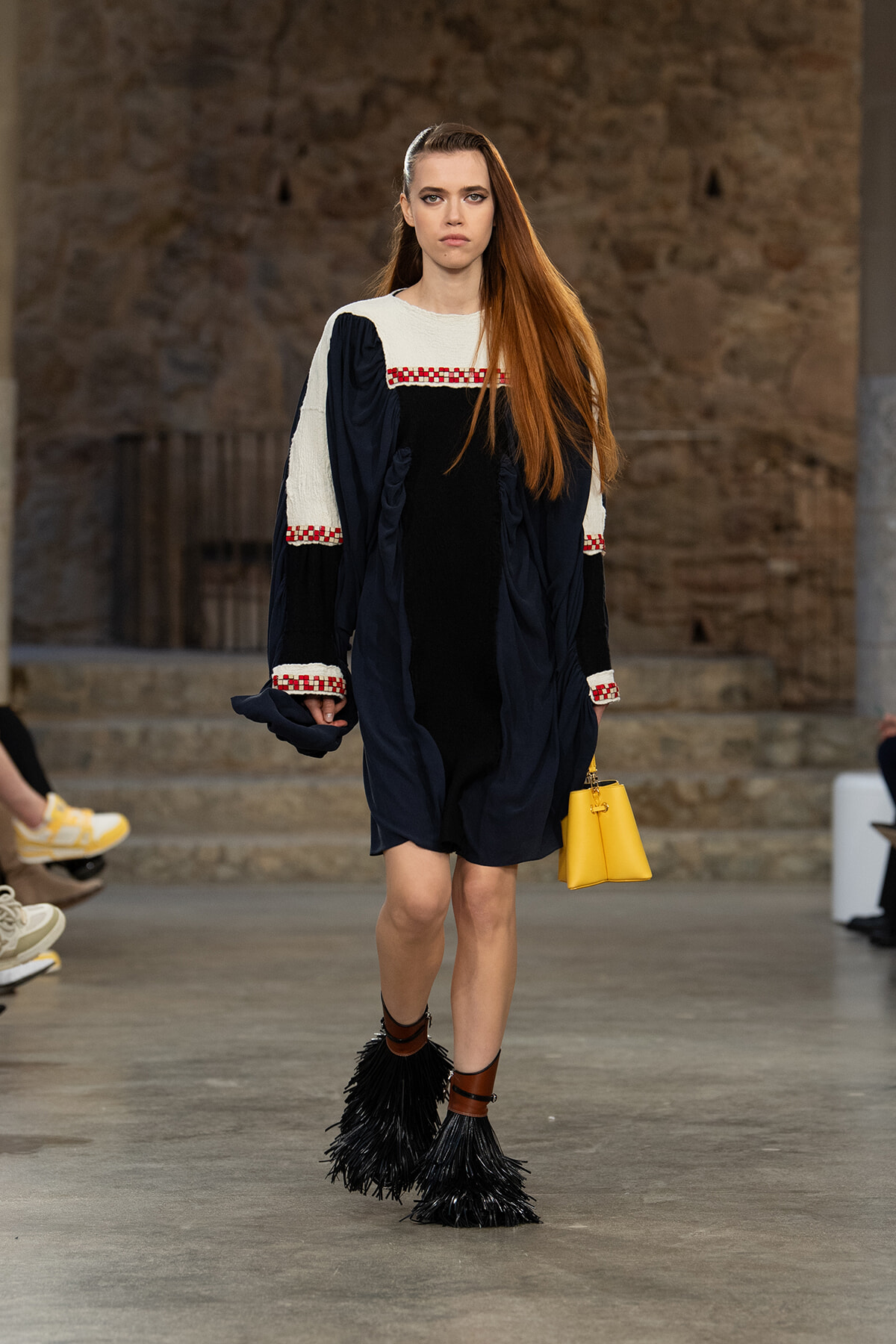 Model walking a runway in a navy dress with white sleeves and red check trim, carrying a yellow handbag and wearing black fringed boots.