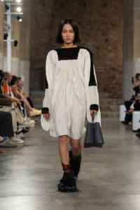 Model on a runway wearing a white, voluminous dress with black yoke and cuffs, carrying a gray handbag and wearing black fringe boots with brown straps.