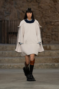 Runway model wearing a loose beige cape-style coat with a dark collar, paired with tall black fringe boots and brown accents.