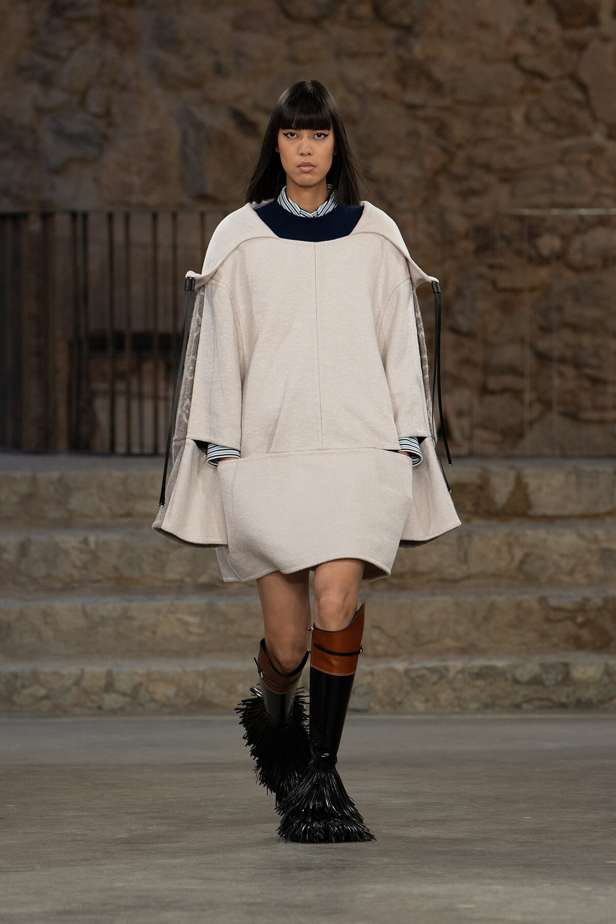 Runway model wearing a loose beige cape-style coat with a dark collar, paired with tall black fringe boots and brown accents.