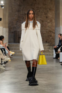 Model walks a runway in an oversized off-the-shoulder white coat over a striped shirt, wearing tall brown-and-black fringe boots and carrying a bright yellow handbag.