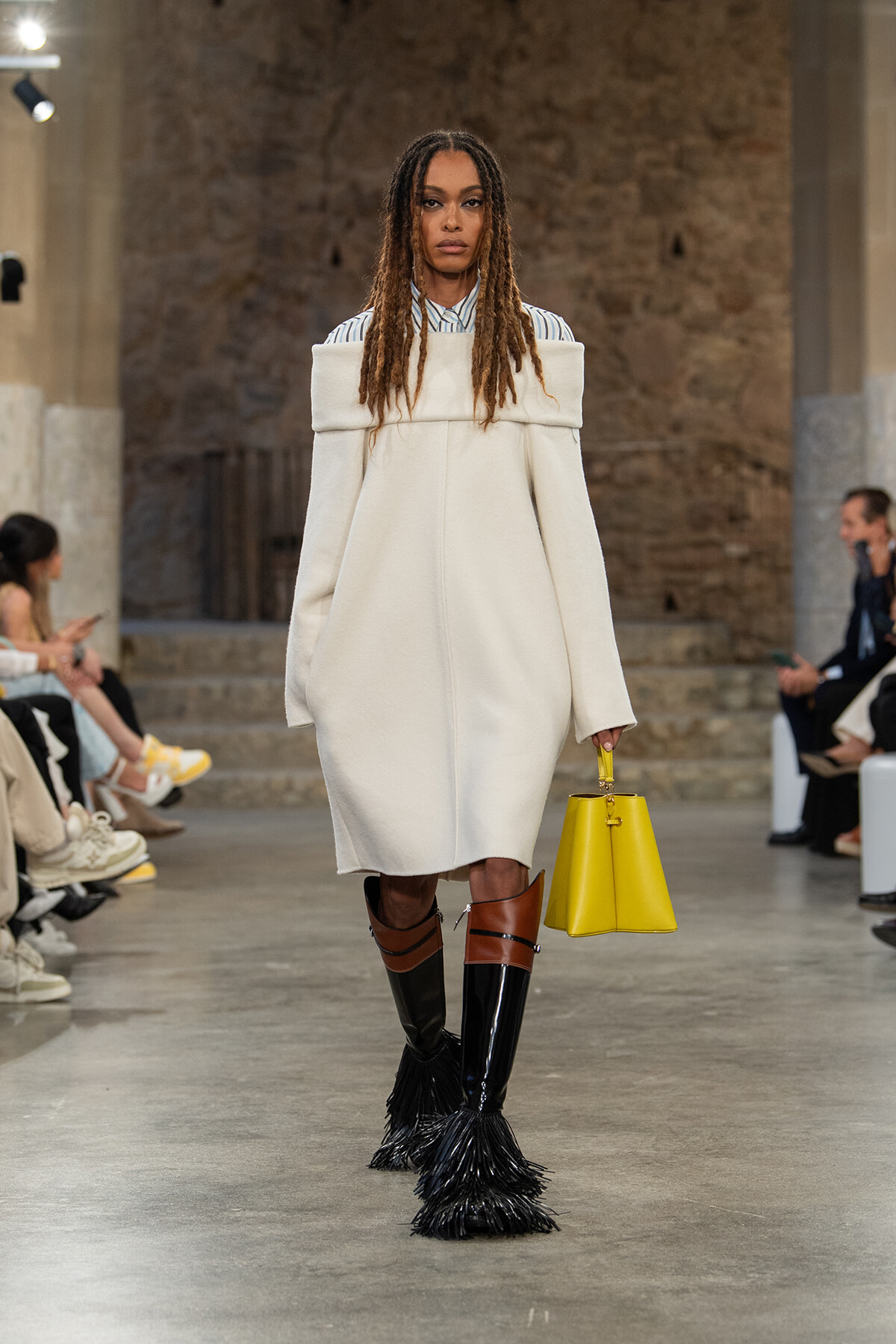Model walks a runway in an oversized off-the-shoulder white coat over a striped shirt, wearing tall brown-and-black fringe boots and carrying a bright yellow handbag.