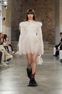 Model walks the runway in a sheer white feathered off-shoulder dress with black fringed boots and a small white bag.
