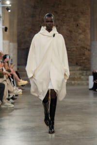 Model walks the runway in a voluminous ivory cape with a large hood and black knee-high boots.