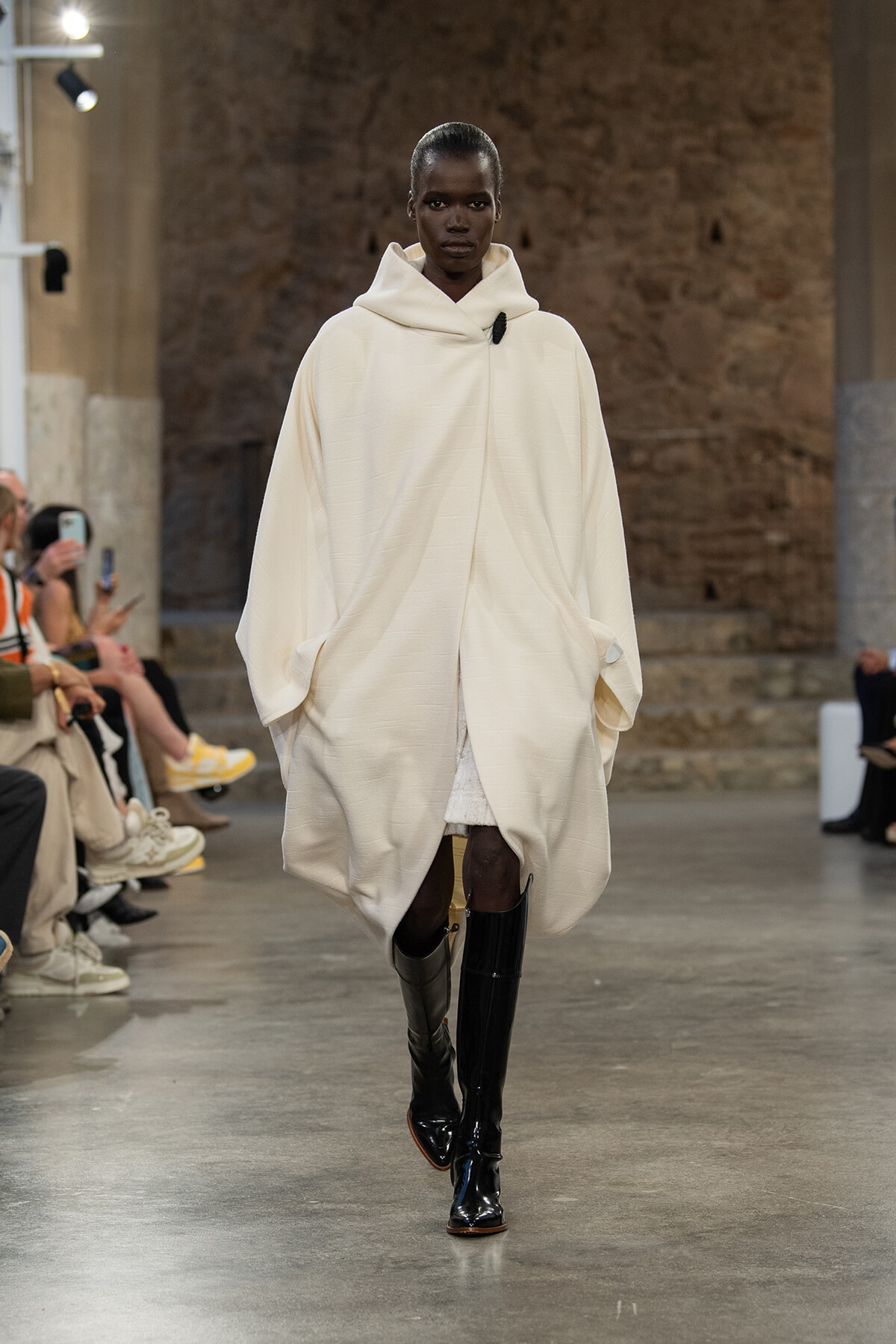 Model walks the runway in a voluminous ivory cape with a large hood and black knee-high boots.
