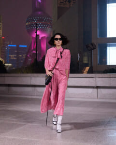 Woman in a pink patterned outfit and sunglasses walks across a city plaza at night, with illuminated buildings in the background.