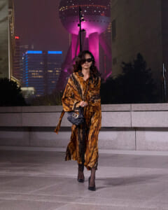 Woman in a tiger-print coat and black sunglasses walking at night on a plaza, carrying a black quilted handbag with a chain strap.