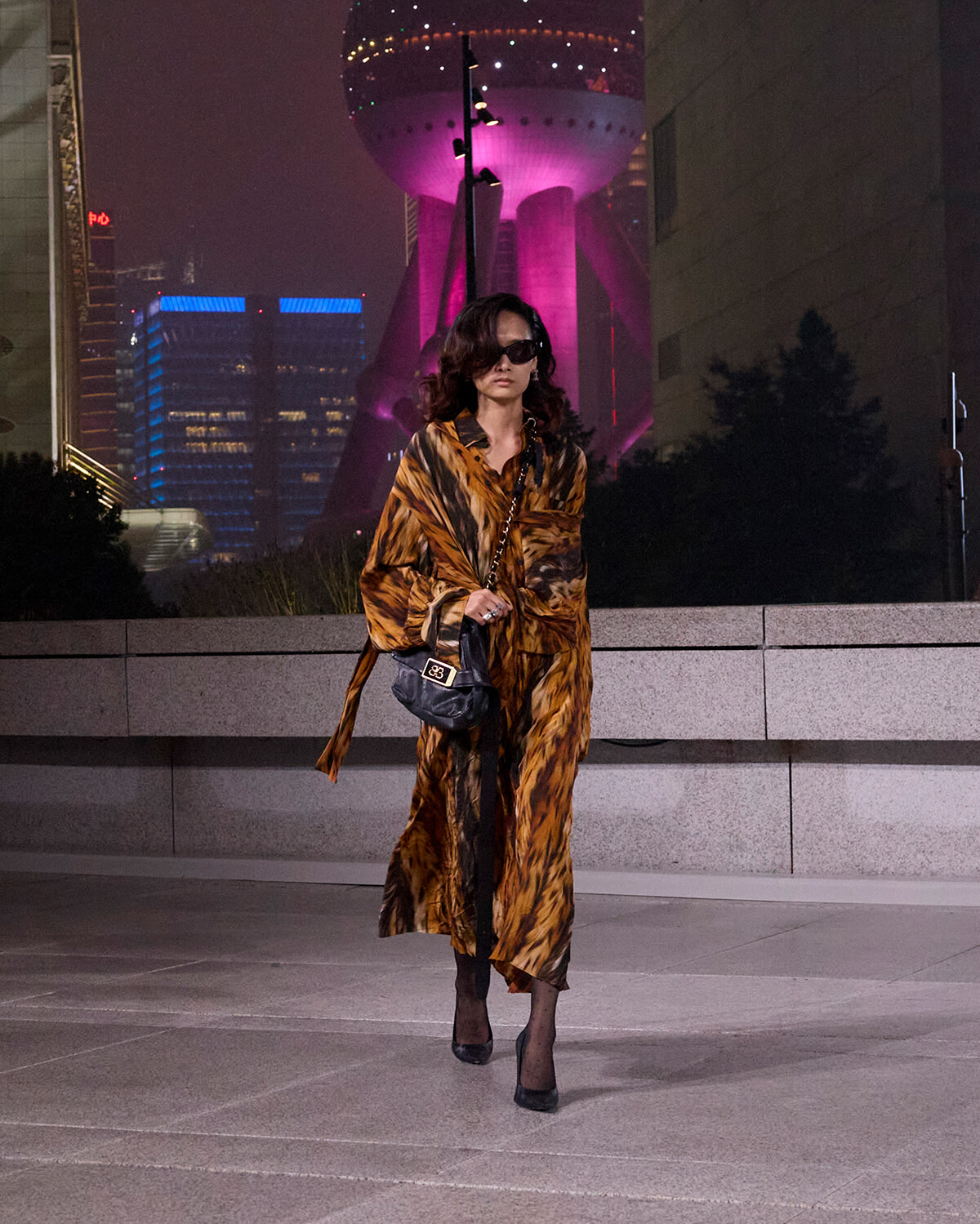 Woman in a tiger-print coat and black sunglasses walking at night on a plaza, carrying a black quilted handbag with a chain strap.