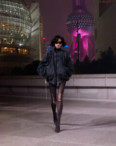 Person in an oversized black bomber jacket and ripped sheer leggings walks at night in a neon city plaza.