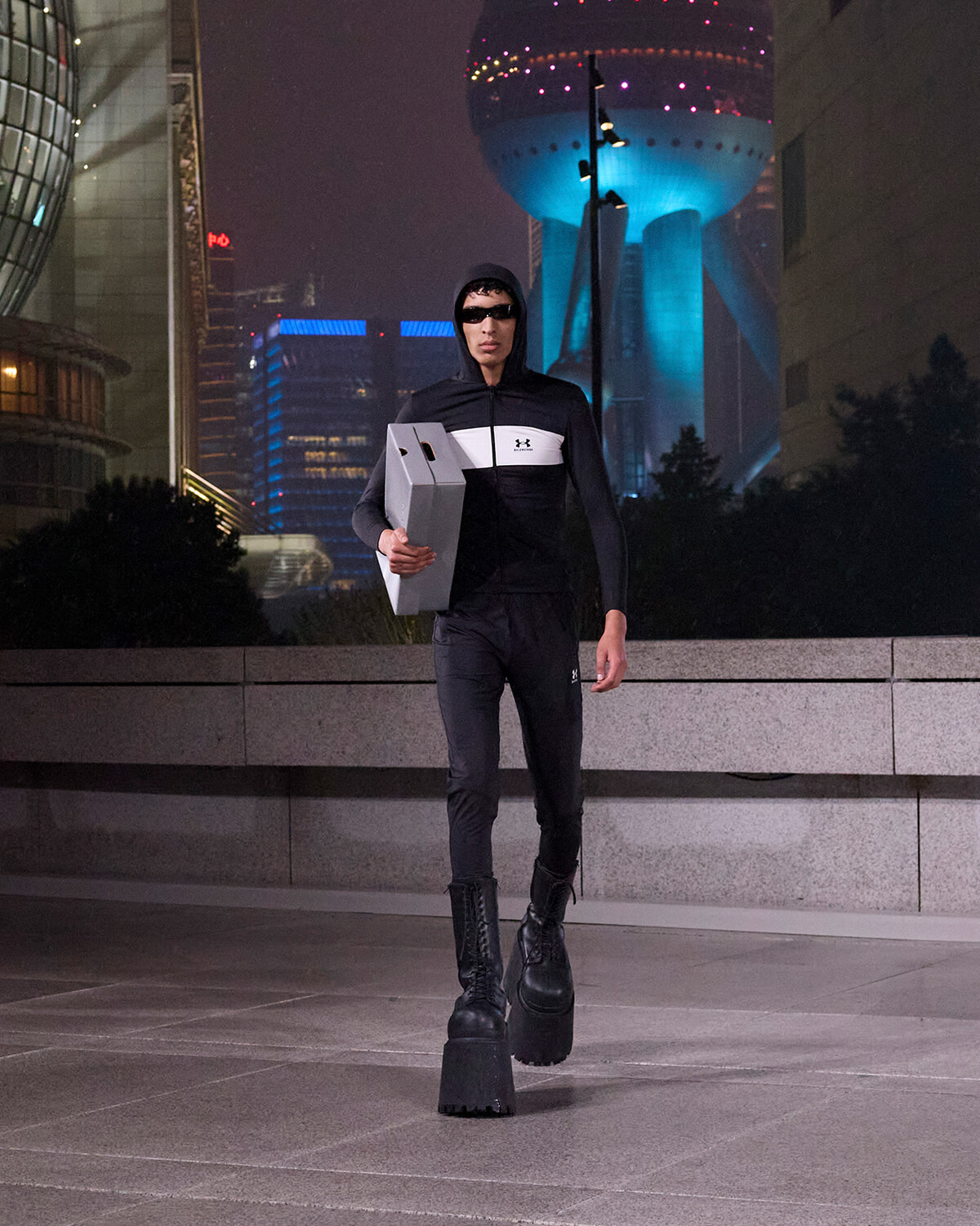 Person in a black hooded jacket and sunglasses walks at night in a futuristic cityscape, carrying a white cardboard box under one arm.