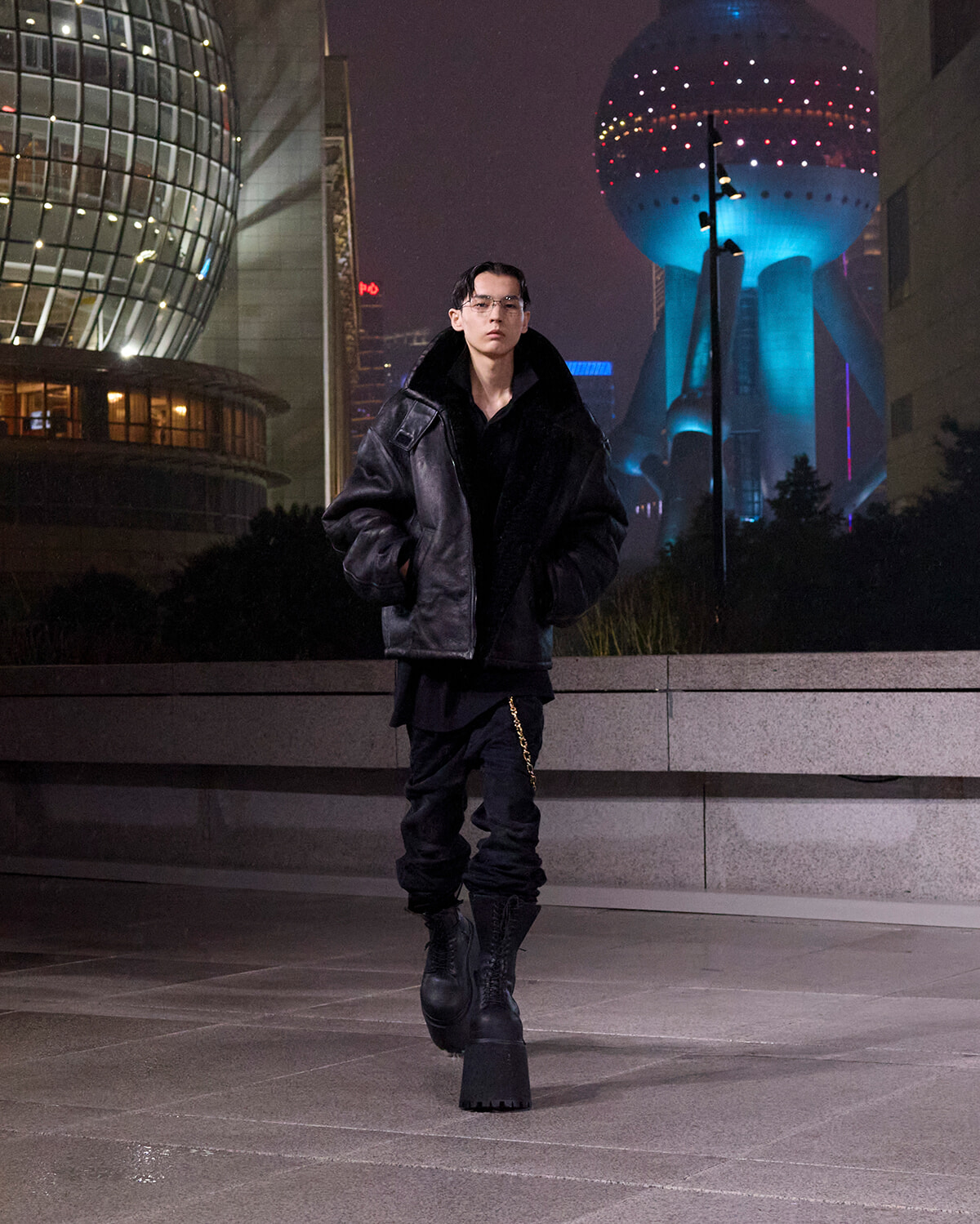 Man in an oversized black leather jacket and chunky boots walks along a city plaza at night, with futuristic buildings lit in the background.
