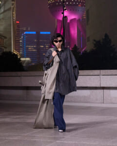 Person in an oversized striped shirt and blue wide-leg pants, carrying a beige trench coat, walking at night with neon city lights behind them.