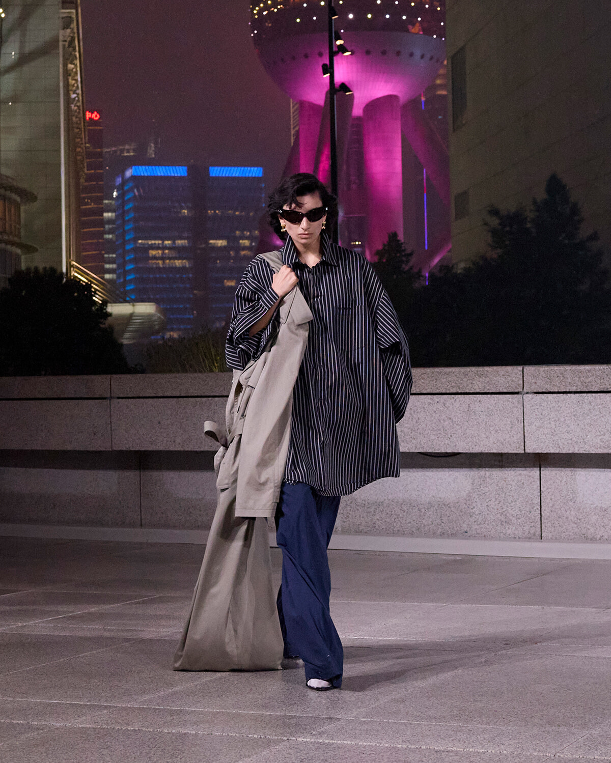 Person in an oversized striped shirt and blue wide-leg pants, carrying a beige trench coat, walking at night with neon city lights behind them.