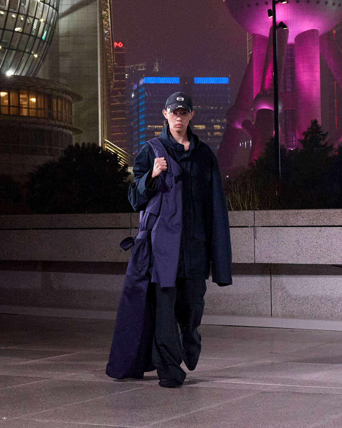 Person wearing a dark oversized coat and cap walks through a nighttime urban plaza, carrying a large purple bag against a cityscape with illuminated buildings and purple lighting in the background.
