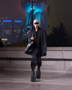 Person wearing an oversized black coat and white cap with visor sunglasses, walking at night in an urban plaza with a large futuristic building in the background, carrying a black bag and a long pole.