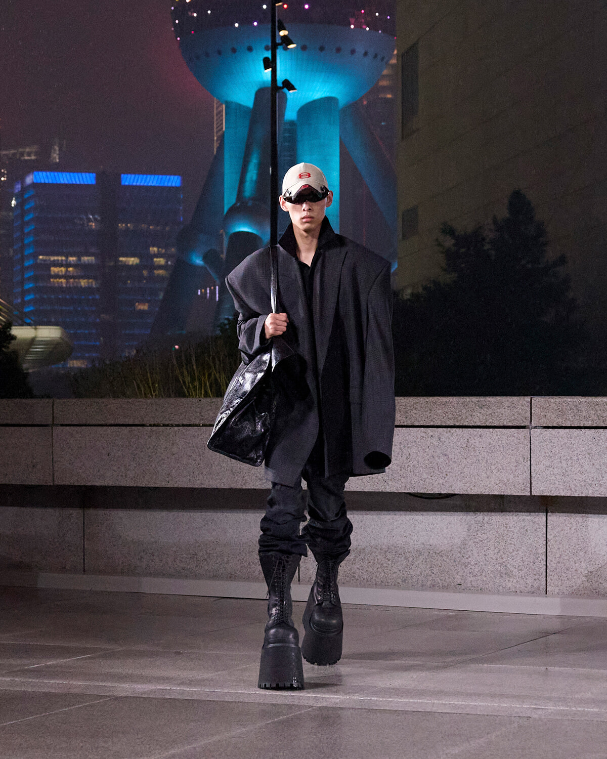 Person wearing an oversized black coat and white cap with visor sunglasses, walking at night in an urban plaza with a large futuristic building in the background, carrying a black bag and a long pole.