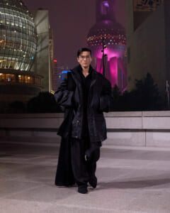 Man in a long black oversized coat walking toward the camera in a nighttime urban plaza with futuristic glass buildings in the background.