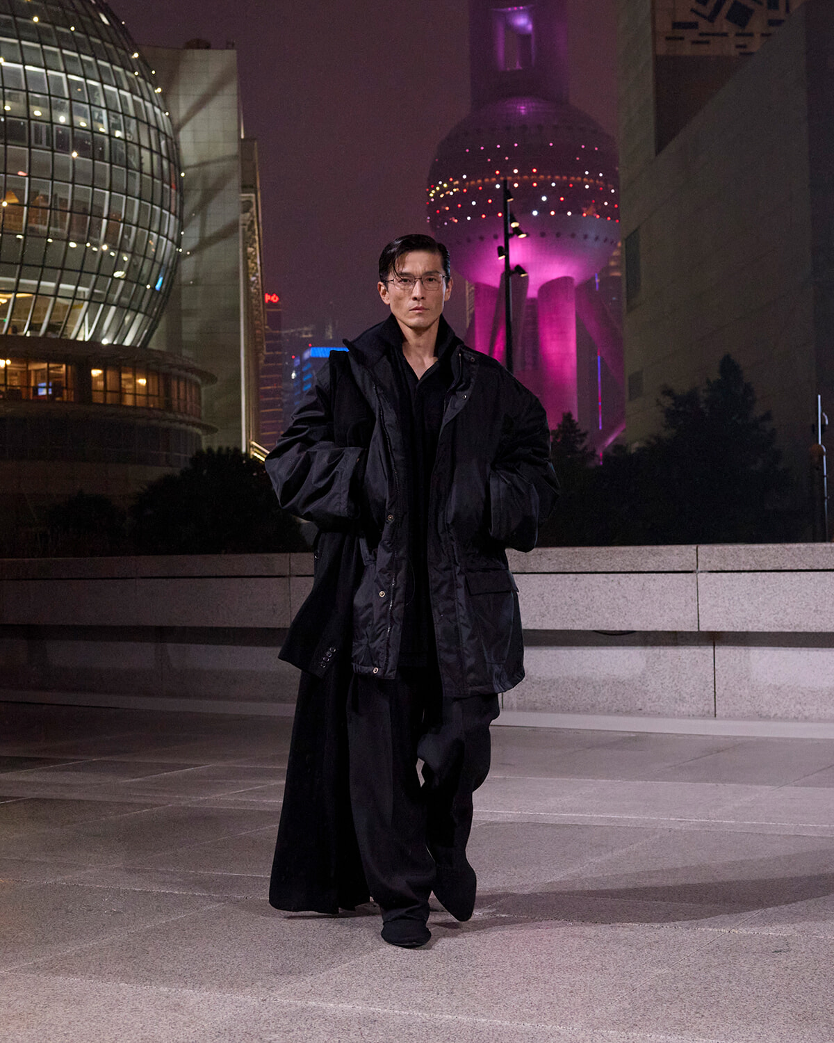 Man in a long black oversized coat walking toward the camera in a nighttime urban plaza with futuristic glass buildings in the background.