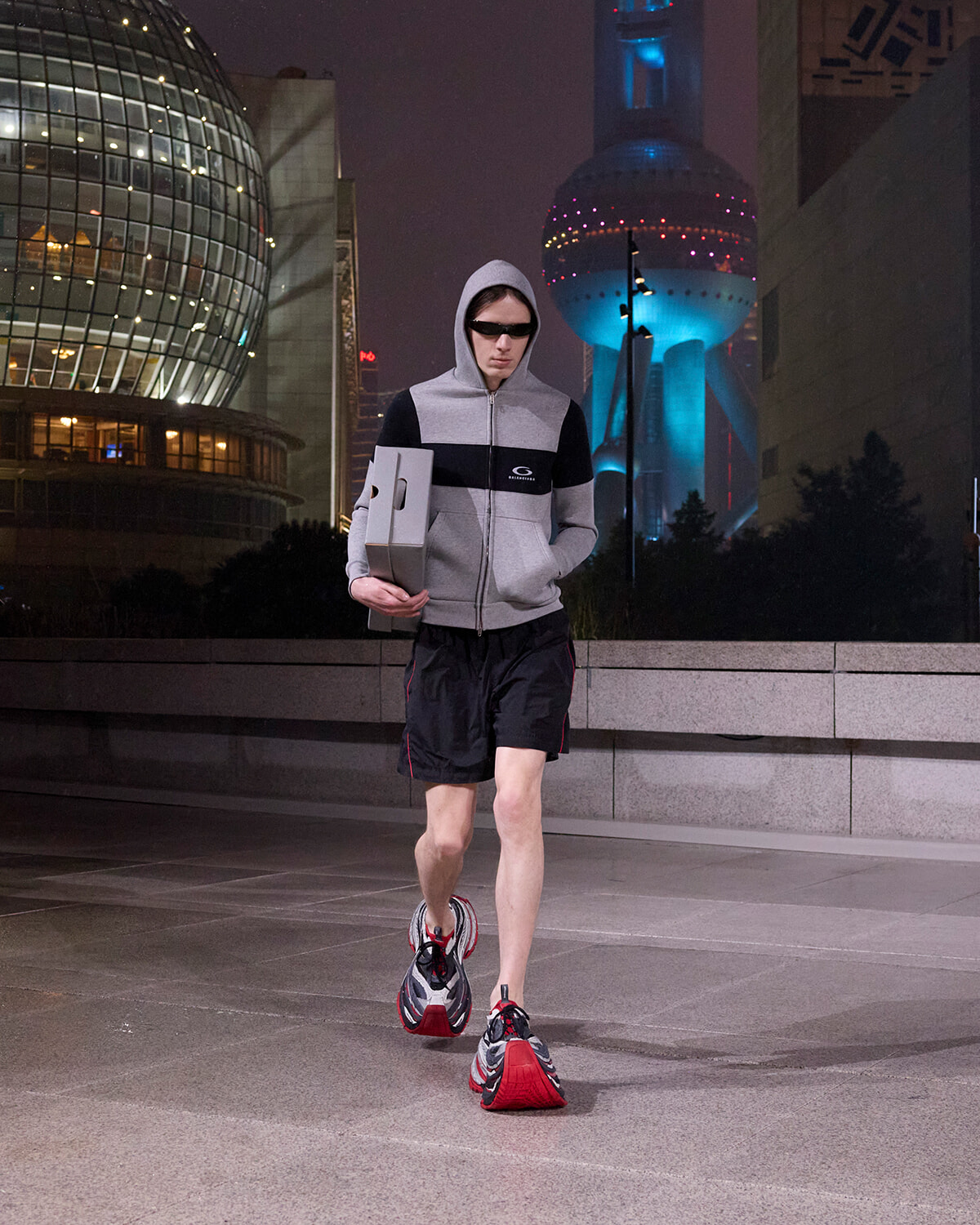 Man in a gray hoodie and black shorts runs at night in a city, carrying two boxes under his arm.