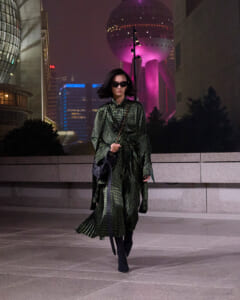 A woman in a green patterned coat walks at night in a city plaza, wearing sunglasses and carrying a black handbag with a chain strap, illuminated skyscrapers behind her.