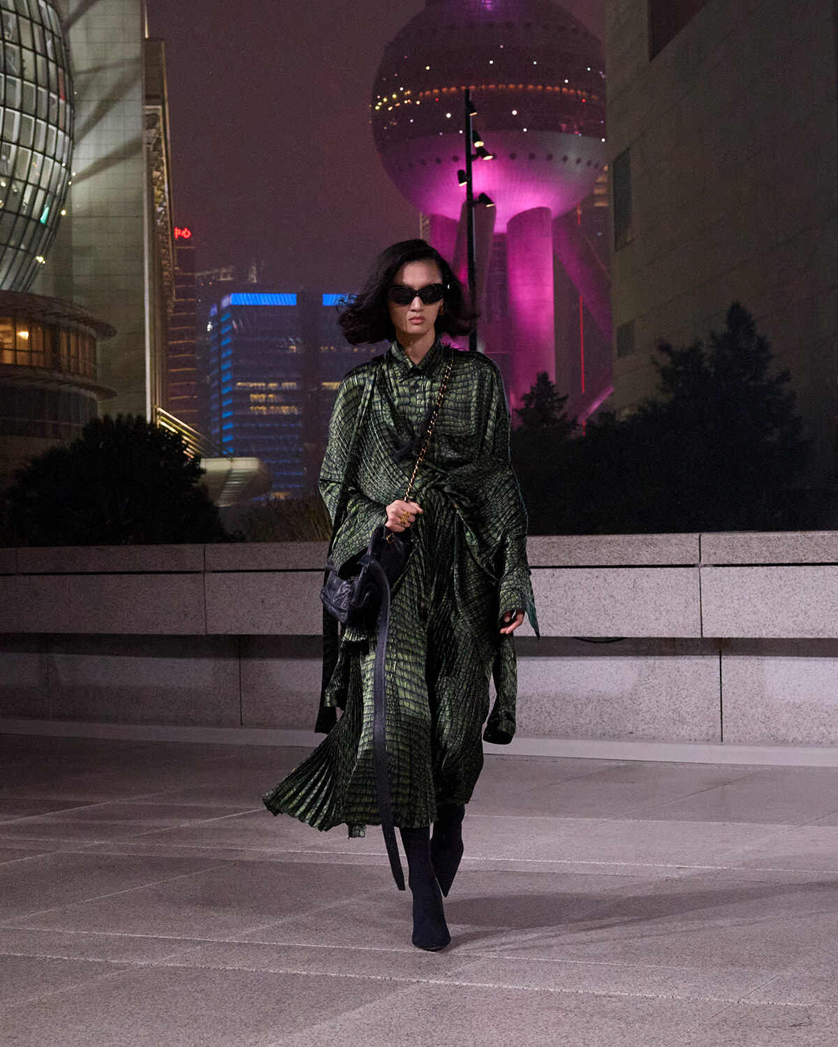 A woman in a green patterned coat walks at night in a city plaza, wearing sunglasses and carrying a black handbag with a chain strap, illuminated skyscrapers behind her.