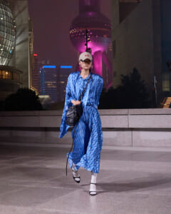 Woman in a blue patterned dress and white cap walks across a nighttime city plaza, carrying a black shoulder bag.