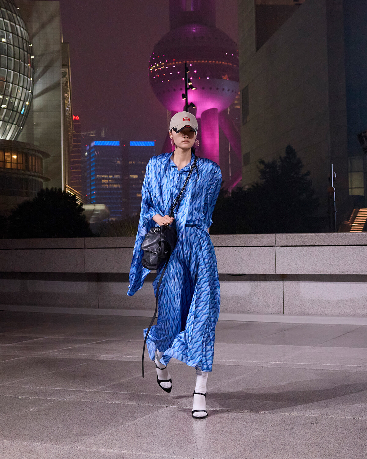 Woman in a blue patterned dress and white cap walks across a nighttime city plaza, carrying a black shoulder bag.