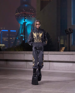 Person standing alone on a city street at night in a black hoodie with gold designs and matching pants, wearing oversized platform boots.