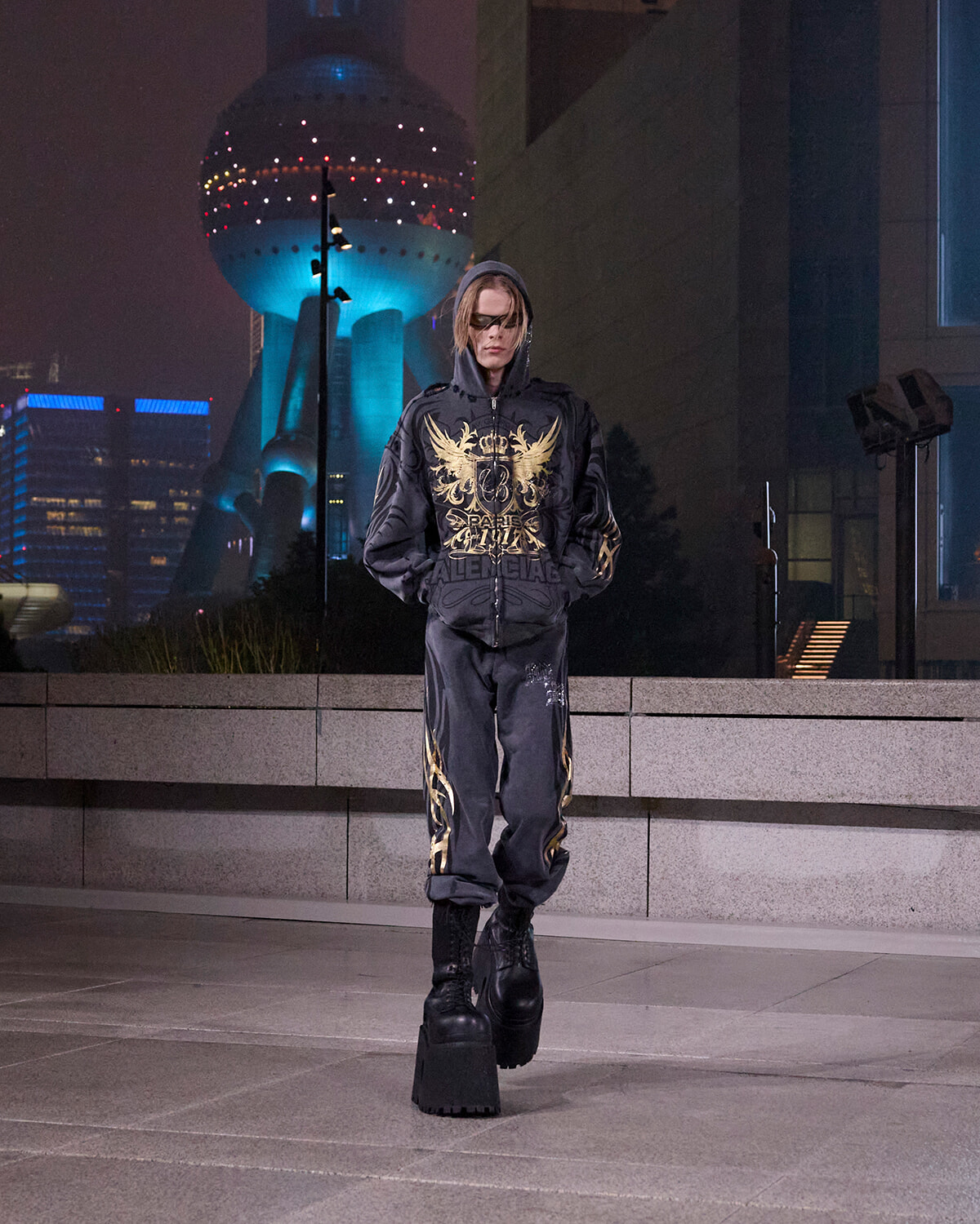 Person standing alone on a city street at night in a black hoodie with gold designs and matching pants, wearing oversized platform boots.
