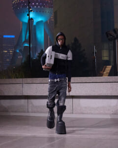 Young man in a hoodie and baggy pants walks at night, carrying two white boxes, with a blue-lit futuristic building in the background.