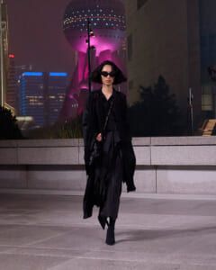 Woman in a flowing black coat and sunglasses walking at night in a neon-lit cityscape.