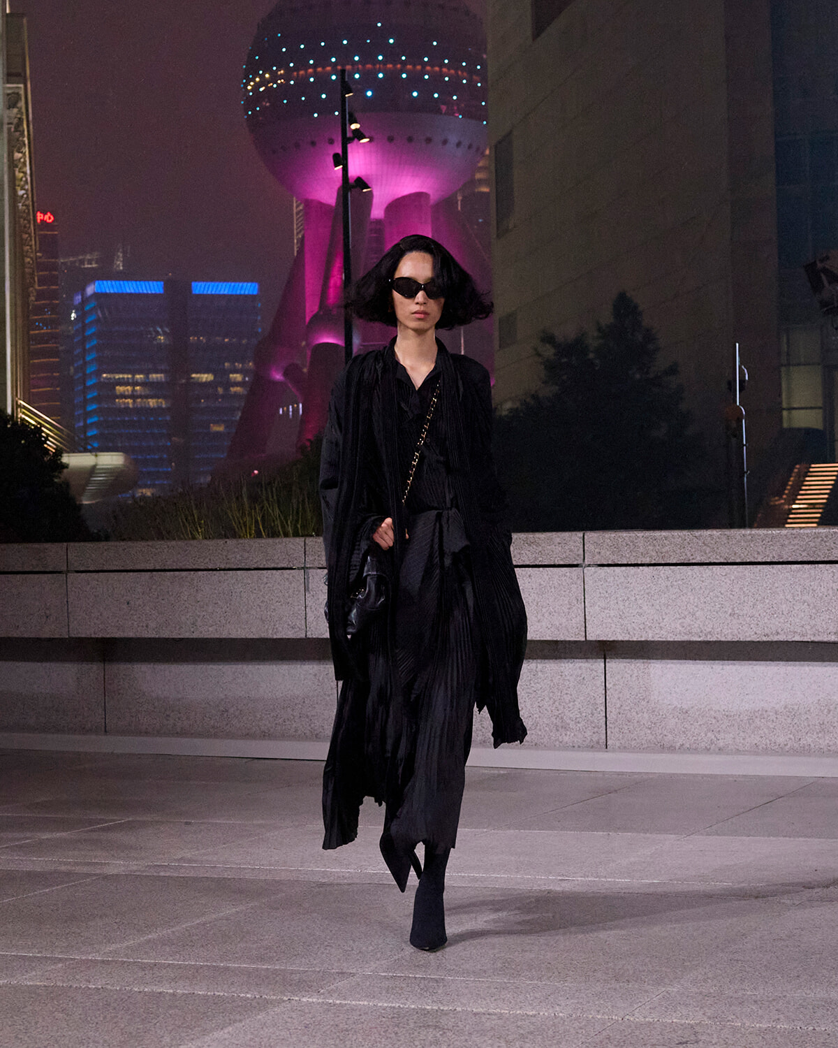 Woman in a flowing black coat and sunglasses walking at night in a neon-lit cityscape.